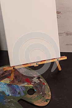 Empty blank white art canvas sitting on painters easel with paint palette