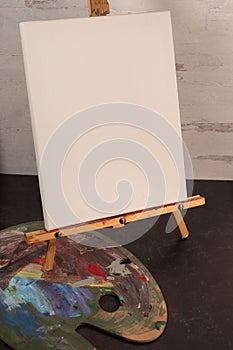 Empty blank white art canvas sitting on painters easel with paint palette