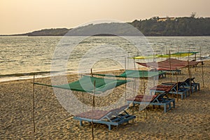 Empty blue beach beds under color awnings on the yellow sand against the background of the sea and the green island with the house