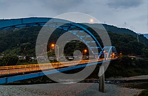 Empty blue bridge in mountain landscape with distant full moon at dusk