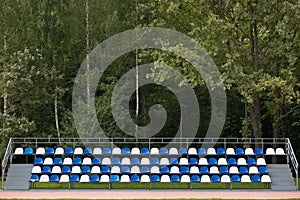 Empty blue and white seats in a football or soccer stadium. Grass field and plastic chairs, open door sports arena