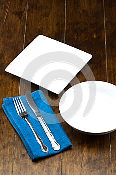 Empty bowl and square dish with fork, knife, napery on wood table.