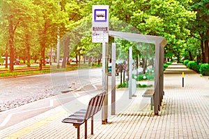 An empty bus stop in the center of the city is lit by the sun, a stop against the background of green trees