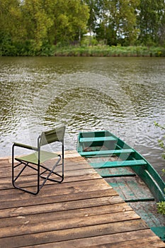 An empty chair and a fishing rod on a wooden pier by the lake, a green boat near the pier. Outdoor recreation.