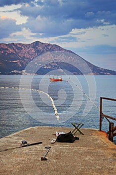 empty chair, fishing rods and fishing gear on the stone pier. in the background of a red boat in the sea