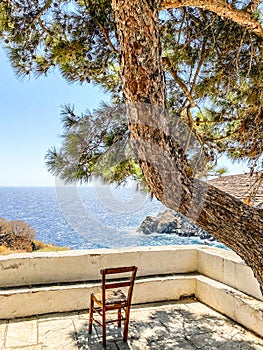 Empty chair on terrace in front of aegean sea under shadow of pine tree, Sifnos island, Greece