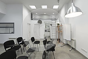 An empty classroom with high ceilings, pendant lamps and lots of black armrest chairs with a swivel table, a white board for