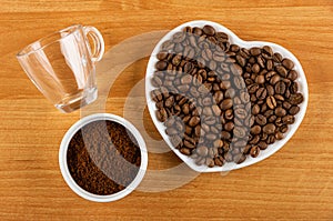 Empty coffeecup, roasted coffee beans in saucer in heart shape, bowl with ground coffee on wooden table. Top view