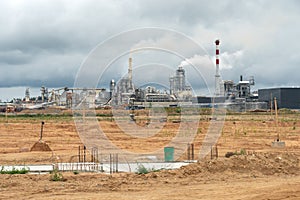 An empty construction site is located next to a large factory. Large factory chimneys emit thick, white, noxious smoke against the