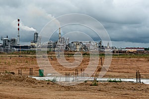 An empty construction site is located next to a large factory. Large factory chimneys emit thick, white, noxious smoke against the