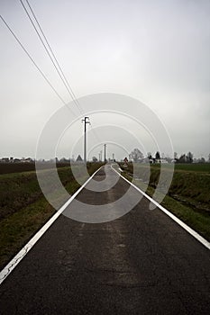 Empty country road bordered by stream of water and electricity pylons on a cloudy day in the italian countryside