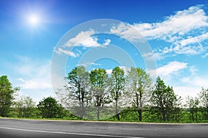 An empty countryside road with trees and bushes against a blue sky with clouds and sun