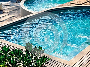 Empty curved swimming pool with clean clear water, nobody. Outdoor pool exterior with splashing water on wood plank decks.