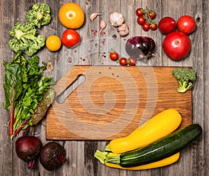 Empty cutting board and vegetables on weathered wooden background. Colorful ingredients for cooking on rustic wooden table around