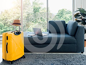 Empty dark sofa seat with passport, laptop computer, and beach hat on yellow travel suitcase on the carpet.