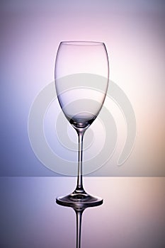 Empty glass goblet without wine on a thin leg stands on a mirror surface. Colorful purple background