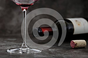 Empty glass of wine in front of lying wine bottle with cork on dark gray background, concept of alcoholism