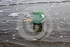 Empty green coffee cup on black sand, washing away by the waves in the sea. Still life concept backgrounds