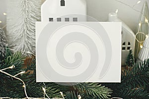 Empty greeting card on background of stylish christmas houses, fir branches with golden lights and tree decorations. Christmas