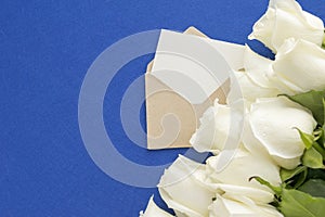 Empty greeting card in open envelope with white rose flowers on blue background. Top angle view. Wedding invitation or