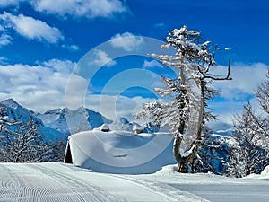 Empty groomed ski resort slope runs past a cottage in Alps covered in fresh snow