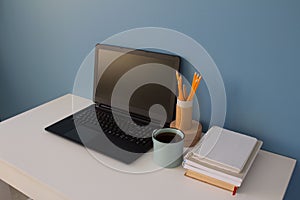 Empty home office of creative entrepreneur with black laptop on white table during lunch break. Modern workspace with blue cup of