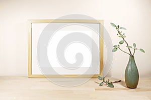 Empty horizontal frame with passe-partout as mock-up and a vase with sage twig on a light wooden table or desk in front of a beige