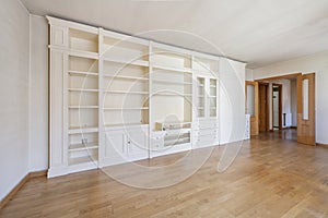 Empty living room with white lacquered wooden bookcase with glass cabinets, many shelves, furniture with drawers and cornices
