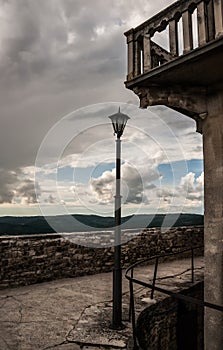 An empty mediterranean street with cracks on the floor, a lamp post and a balcony under a cloudy sky.