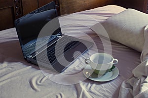 An empty mug on a dirty saucer next to an open laptop on an unmade bedroom bed with rumpled blankets.