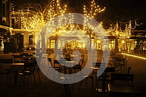 Empty night restaurant, lot of tables and chairs with noone, magic fairy lights on trees like christmas celebration photo