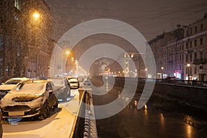 Empty night winter road with cars parked along street during snowfall. Evening city in snowstorm