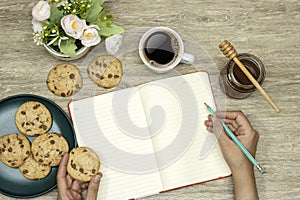 empty notebook, hand holding a pencil an cookies, cup coffee with homemade chocolate chip cookies with honey jar on wood table