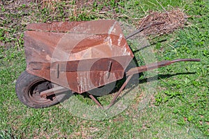 Empty old rusty iron wheelbarrow with one front wheel in rural garden on background of greenery close up, side view