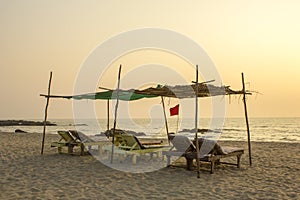 A empty old wooden beach loungers under a palm canopy on the sandy beach of the ocean in the evening. red flag on the sea coast
