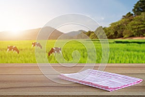 Empty old wooden table and pink checked tablecloth over blurred background of the rural meadows and cows, mountain, blue sky among