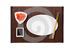 Empty oval white plate with chopsticks for sushi, ginger and soy sauce on dark bamboo mat background. Top view with copy space for