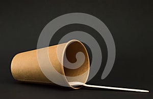 Empty paper cup for coffee lies on its side on a dark background, next to it is a wooden coffee spoon