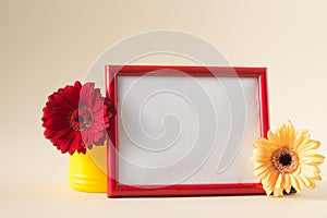 Empty photo frame with red and yellow gerbera daisy flowers on pastel beige background. Picture frame mockup with flowers, Copy