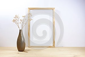 Empty picture frame mockup and a brown ceramic vase with filigree gypsophila on a wooden table against a light gray wall, minimal