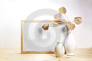 Empty picture frame and two ceramic vases, dry eucalyptus branch with reddish leaves, minimal still life on a wooden table against