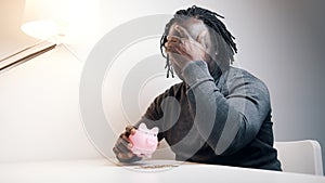 Empty piggy bank. Sad african american man putting one coin inside. Small savings
