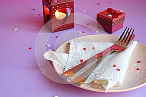 Empty pink plate, cutlery, hearts, candlesticks and red gift on pink-purple background. St. Valentine`s Day table setting concept