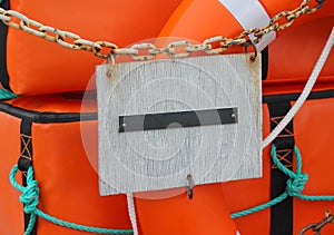 An empty plate attached to an old rusty chain in front of an orange life buoy and an orange boat safety kit