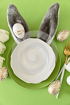 Empty plate decorated bunny ears, white tulips and Easter eggs on green background. Easter celebration concept. Flat lay. Top view