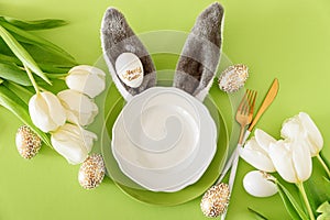 Empty plate decorated bunny ears, white tulips and Easter eggs on green background. Easter celebration concept. Flat lay. Top view