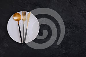 Empty plate with fork, knife and spoon on dark stone background. Gold and black tableware with white plate