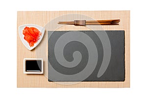 Empty rectangular black slate plate with chopsticks for sushi, ginger and soy sauce on brown sushi mat background. Top view with