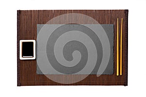 Empty rectangular black slate plate with chopsticks for sushi and soy sauce on dark bamboo mat background. Top view with copy