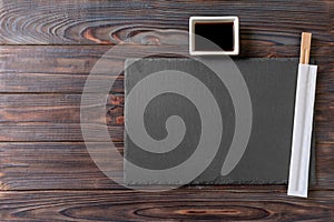Empty rectangular black slate plate with chopsticks for sushi and soy sauce on wooden background. Top view with copy space